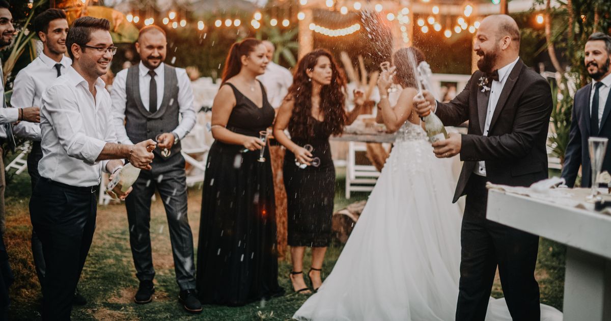 Zdjęcie przedstawia wesele w plenerze, podczas którego pan młody i światek otwierają butelki szampana.