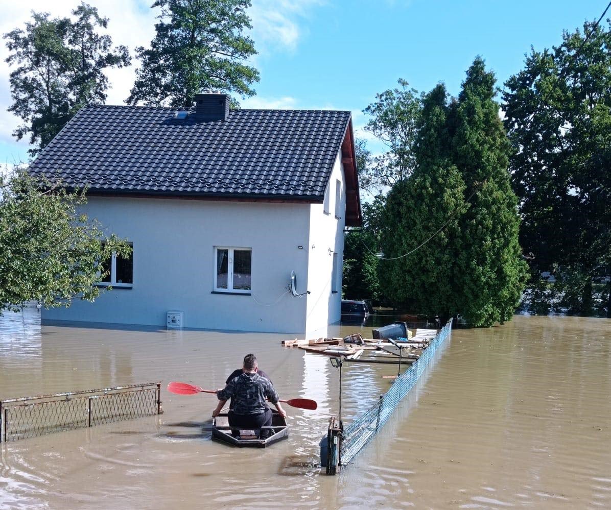 Dwóch mężczyzn płynie kajakiem w stronę podtopionego domu jednorodzinnego.